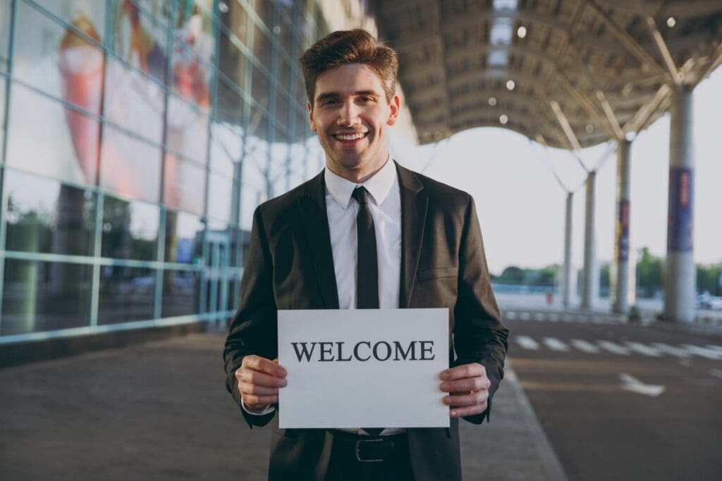 Friendly young satisfied traveler businessman man 20s in black suit stand outside at international airport terminal hold card sign with welcome title text waving hand Air flight business trip concept.